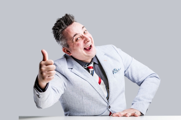 A white man with greying hair light grey suit gives a thumbs up to the camera while posing in a comedic manner